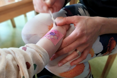 Dieses Foto zeigt, wie eine pädagosische Fachkraft einem Kind ein buntes Glitzer-Tattoo auf dem Arm malt.