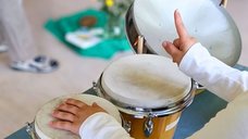 Dieses Bild zeigt eine Nahaufnahme von einem Kind, das mit den Händen eine Percussions-Instrument spielt. Der Zeigefinger der rechten Hand ist in die Luft gestreckt, um den Takt anzugeben.