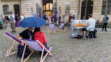 Das Bild zeigt zwei Personen die in einer Fußgängerzone auf Liegestühlen Sitzen. In der Hand hält eine Person einen Regenschirm. Etwas entfernt sitzt ein Mann am Klavier. 