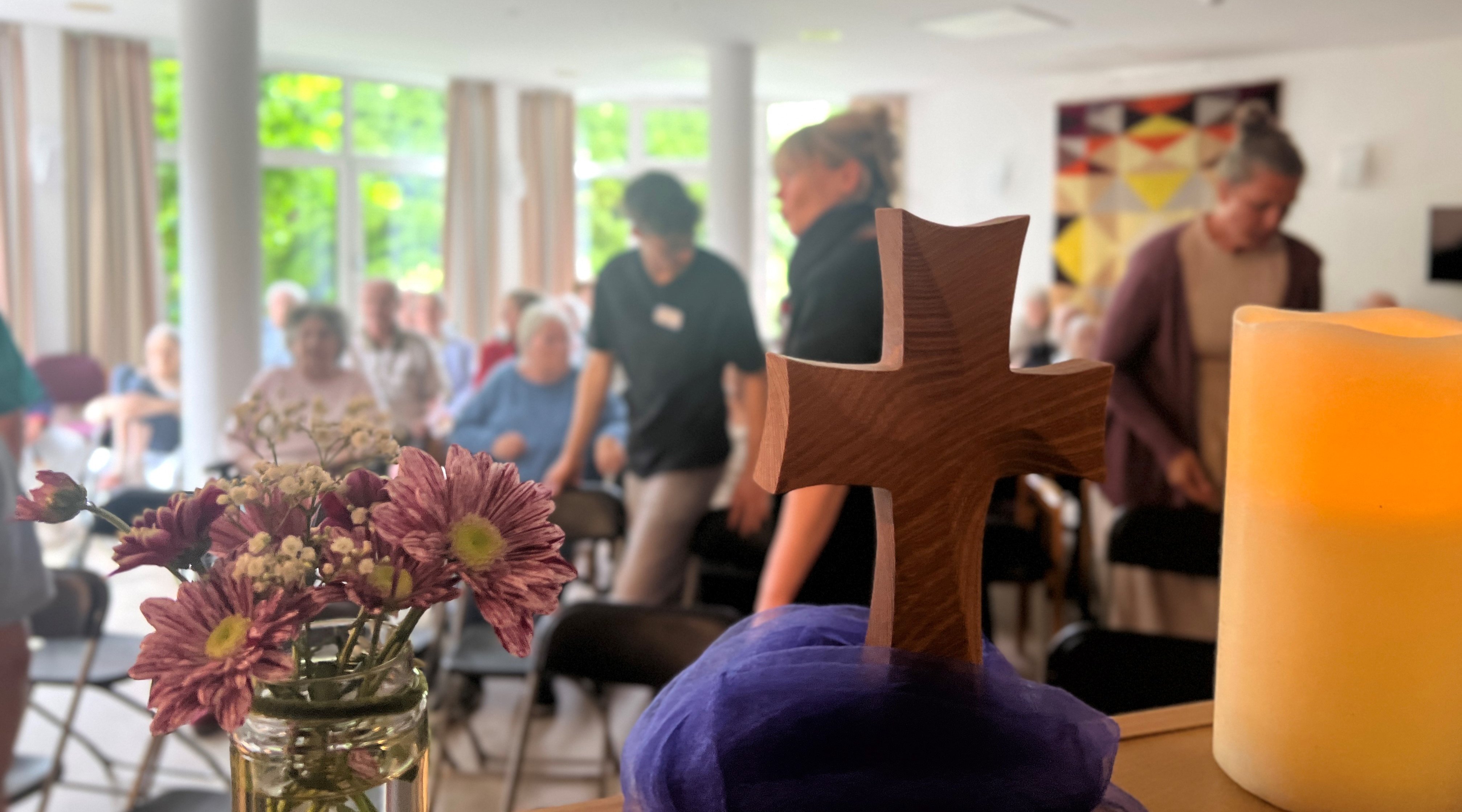 Das Foto zeigt ein Holzkreuz, Blumen und eine brennende Kerze. Im Hintergrund sind Menschen zu sehen, die gerade ihre Plätze einnehmen.