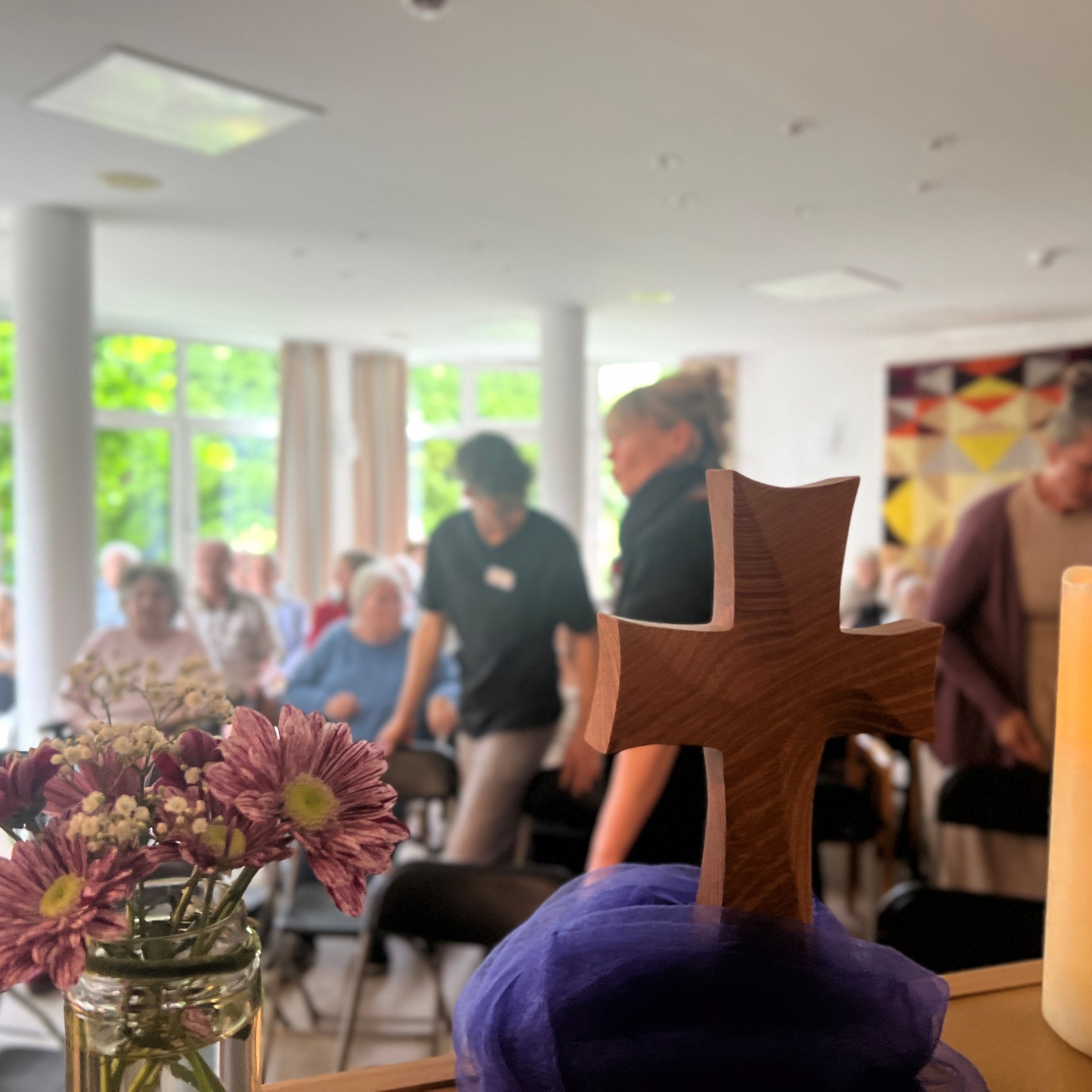 Das Foto zeigt ein Holzkreuz, Blumen und eine brennende Kerze. Im Hintergrund sind Menschen zu sehen, die gerade ihre Plätze einnehmen.