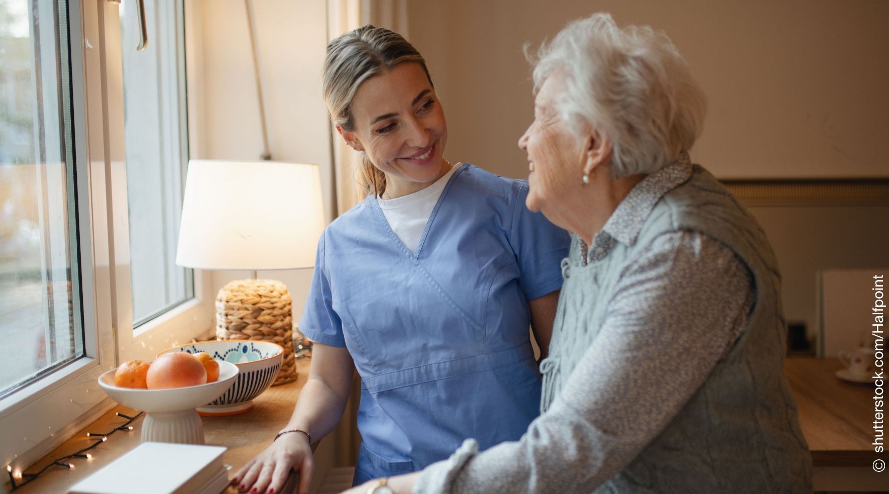 Eine Pflegerin steht mit einer älteren Dame am Fenster und lächelt Sie an.