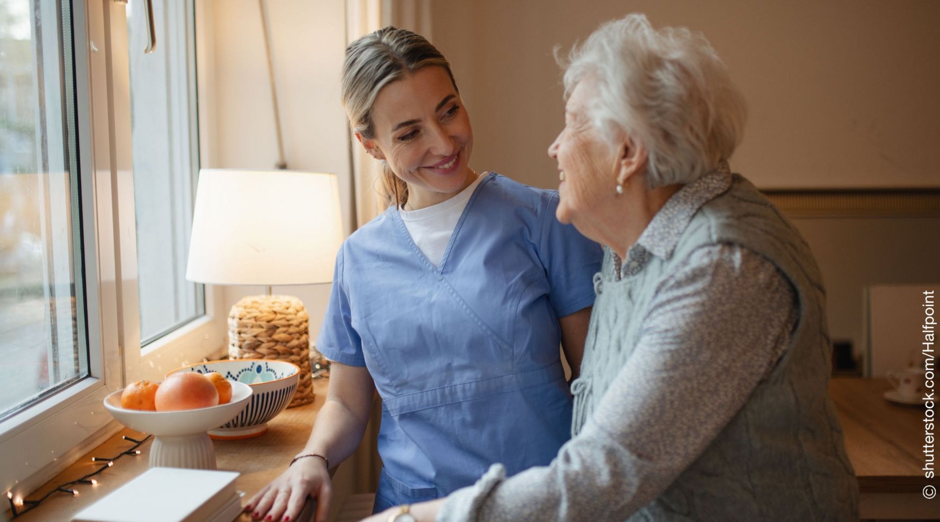 Eine Pflegerin steht mit einer älteren Dame am Fenster und lächelt Sie an.