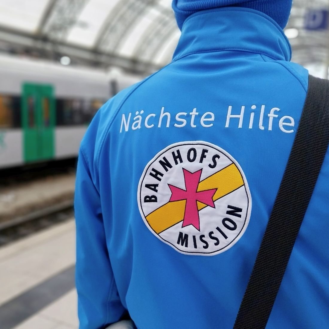 Das Bild zeigt einen ehrenamtlichen Mitarbeiter der Bahnhofsmission Dresden von hinten, der am Bahnsteig unterwegs ist und Ausschau hält, ob irgendwo Hilfe benötigt wird.