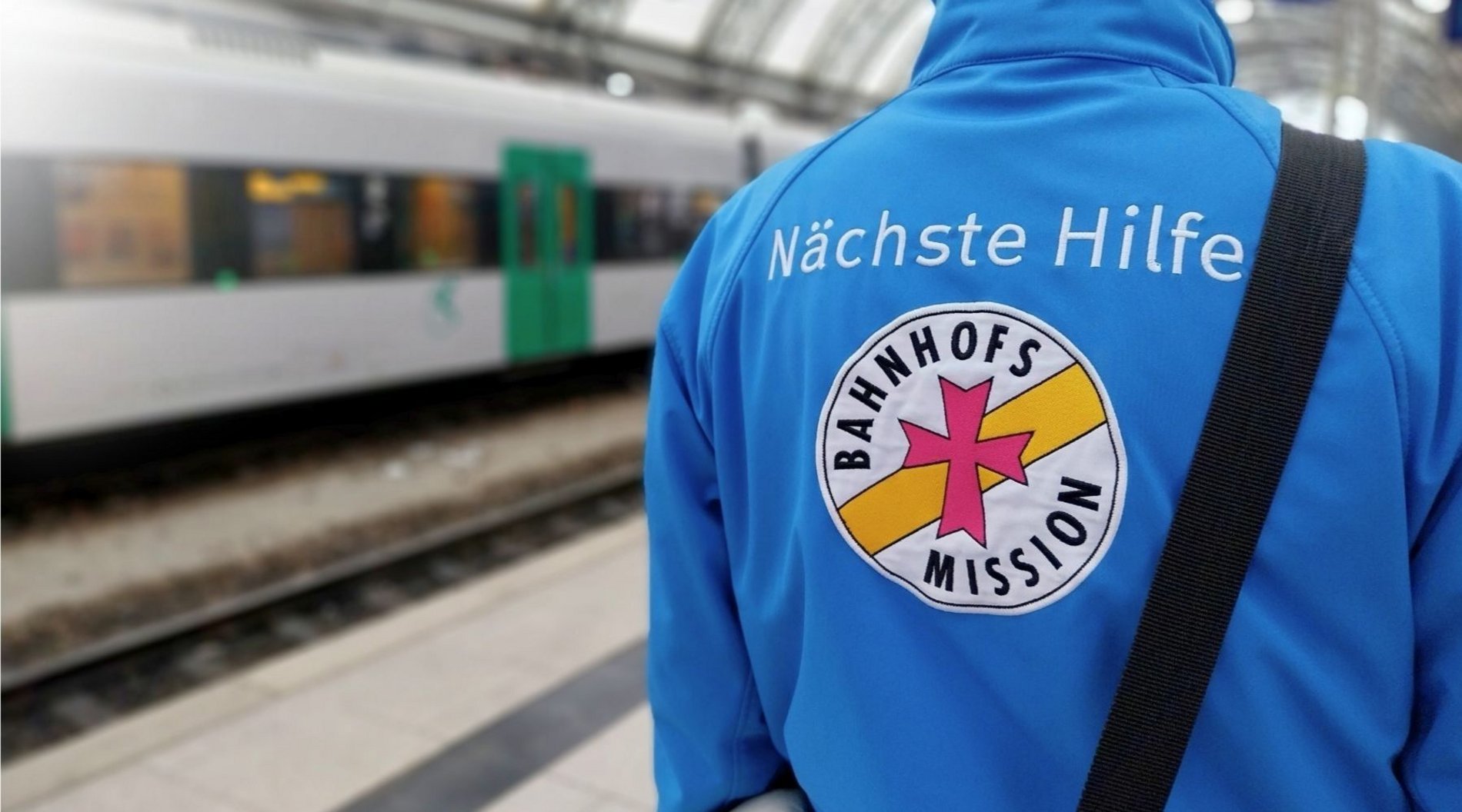 Auf diesem Bild steht ein ehrenamtlicher der Bahnhofsmission Dresden in blauer Bahnhofsmissions-Jacke am Gleis am Dresdner Hauptbahnhof. Die Person ist von hinten zu sehen. Auf der Jacke ist auf dem Rücken das große Logo der Bahnhofsmission zu sehen. Im Hintergrund fährt ein Zug ein.