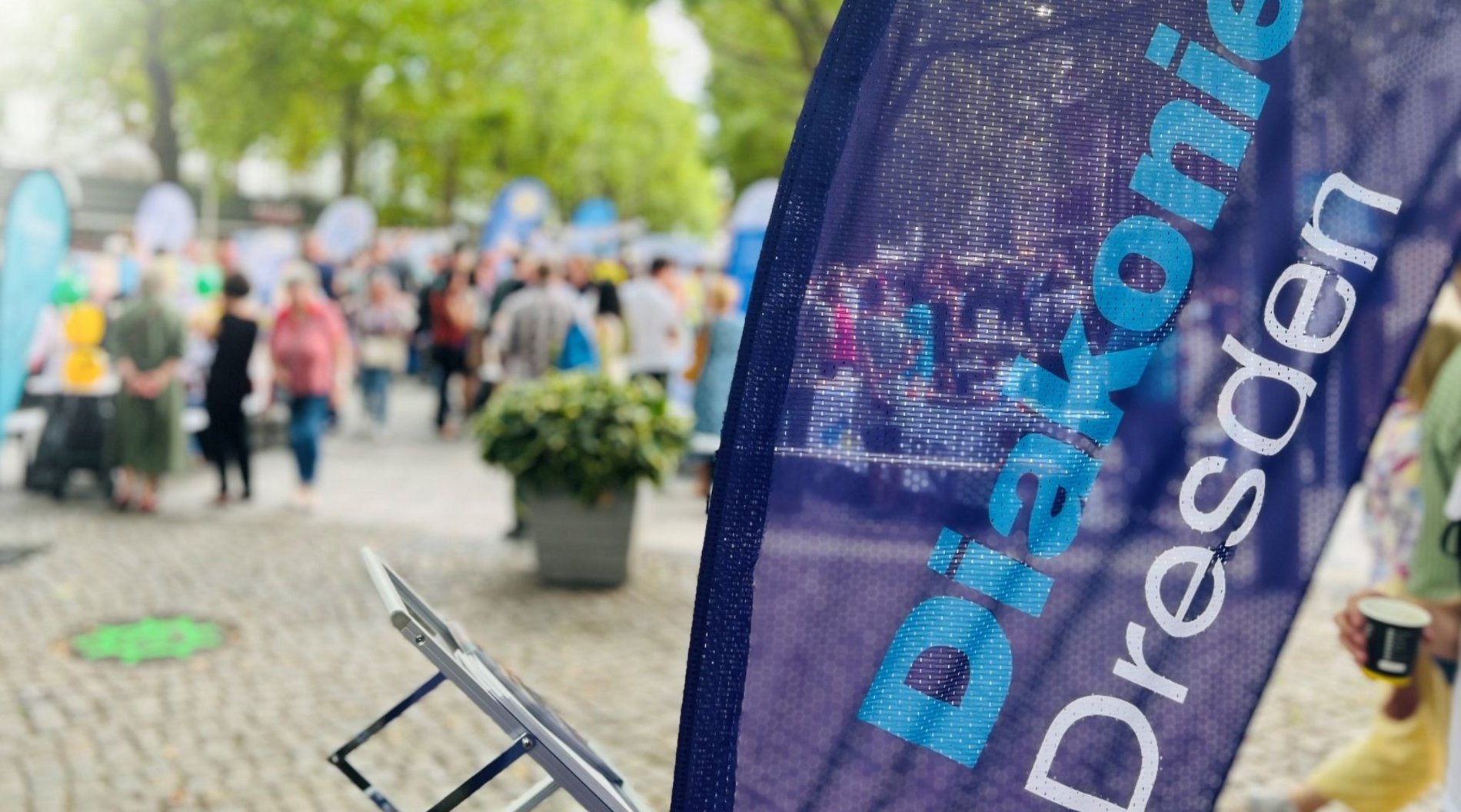 Dieses Bild zeigt einen Ausschnitt des Gastmahls im vorausgegangenen Jahr: Im Vordergrund ist die Beachflag der Diakonie Dresden zu sehen, im Hintergrund tummeln sich Menschen entlang der Tischmeile.