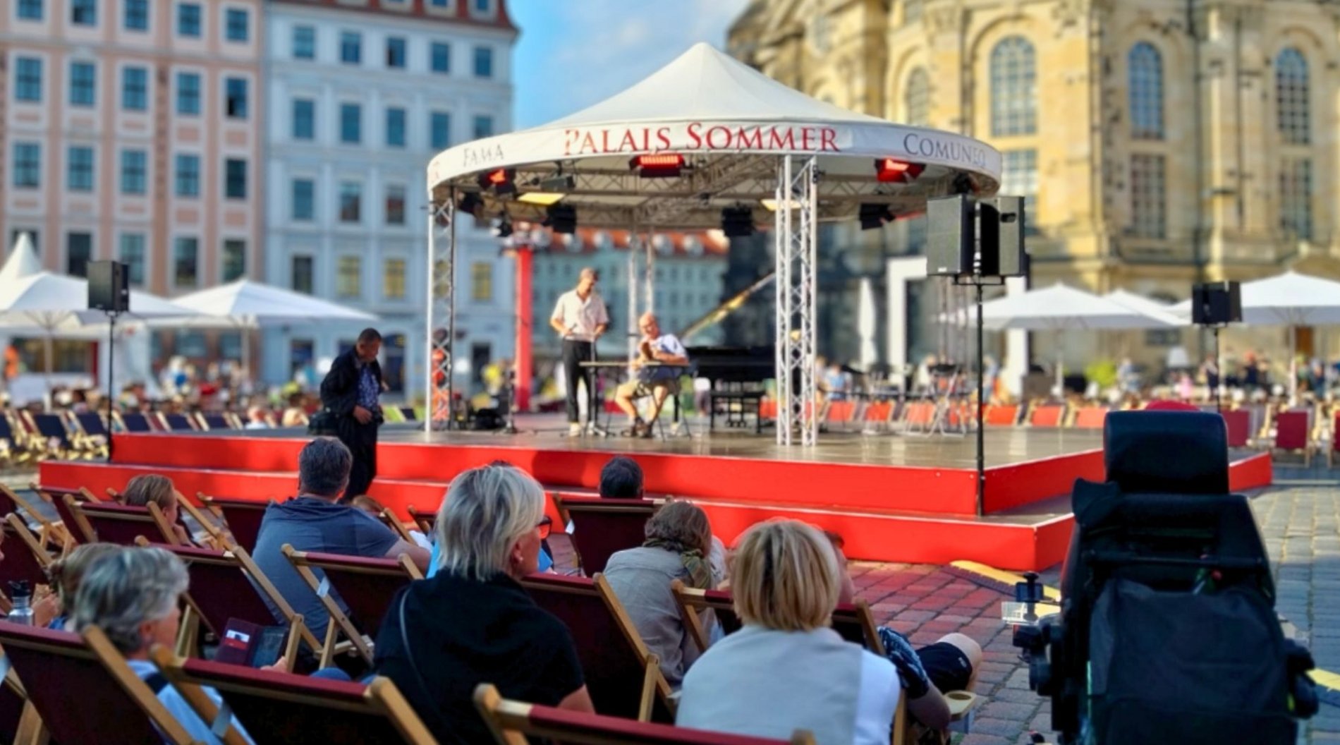 Dieses Bild zeigt die Bühne des Palais Sommers auf dem Neumarkt beim Auftritt der Lesebühne des ABZ im vergangenen Jahr. Im Vordergrund ist das Publikum von hinten zu sehen, ganz hinten sieht man den unteren Teil der Dresdner Frauenkirche.