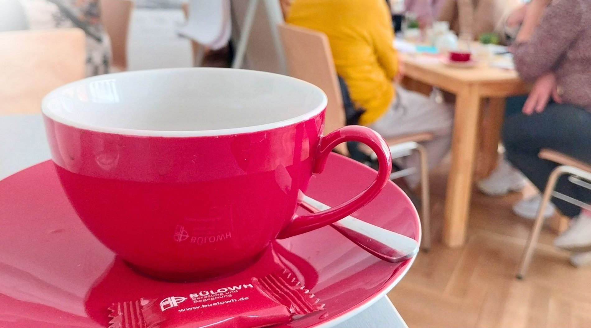 Das Bild zeigt eine Nahaufnahme einer roten Tasse beim Sprachcafé für zugewanderte Menschen 60+ im HAUS DER BRÜCKE. Im Hintergrund sieht man verschwommen die Teilnehmenden.