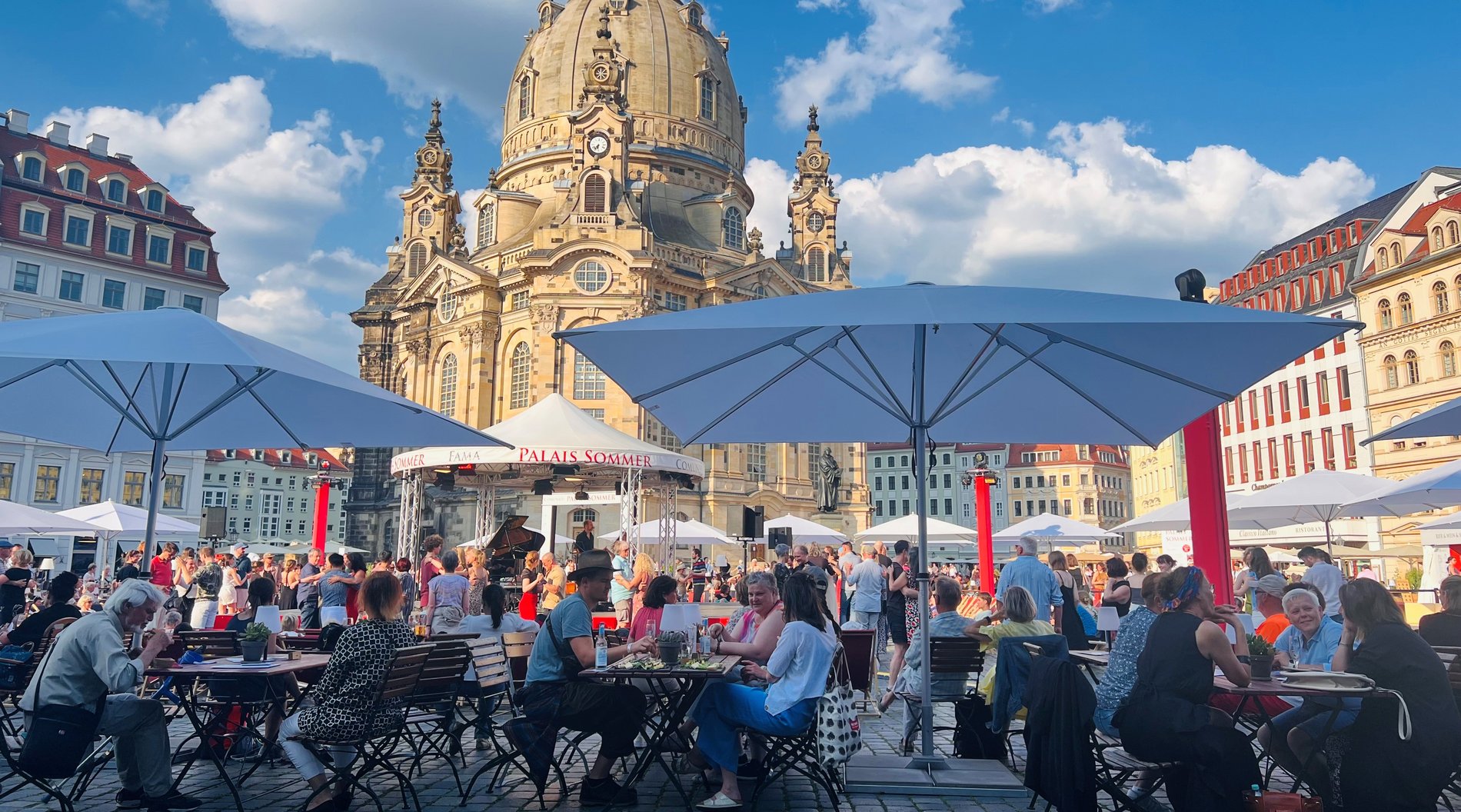 Das Bild zeigt das Festivalgelände auf dem den Neumarkt. Viele Menschen sitzen an Tischen unter Sonnenschirmen. Sie schauen de mbunten Treiben auf der Palais-Sommer-Bühne zu. Im HIntergrund ist die Frauenkirche zu sehen