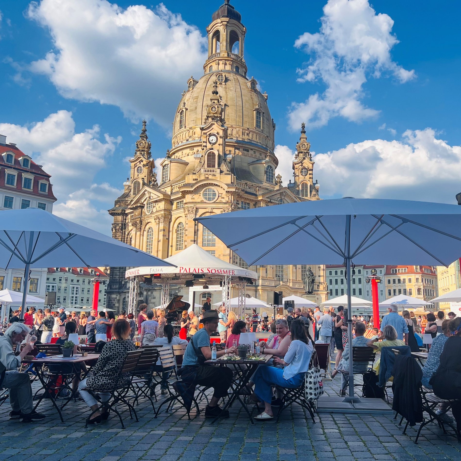 Das Bild zeigt das Festivalgelände auf dem den Neumarkt. Viele Menschen sitzen an Tischen unter Sonnenschirmen. Sie schauen de mbunten Treiben auf der Palais-Sommer-Bühne zu. Im HIntergrund ist die Frauenkirche zu sehen