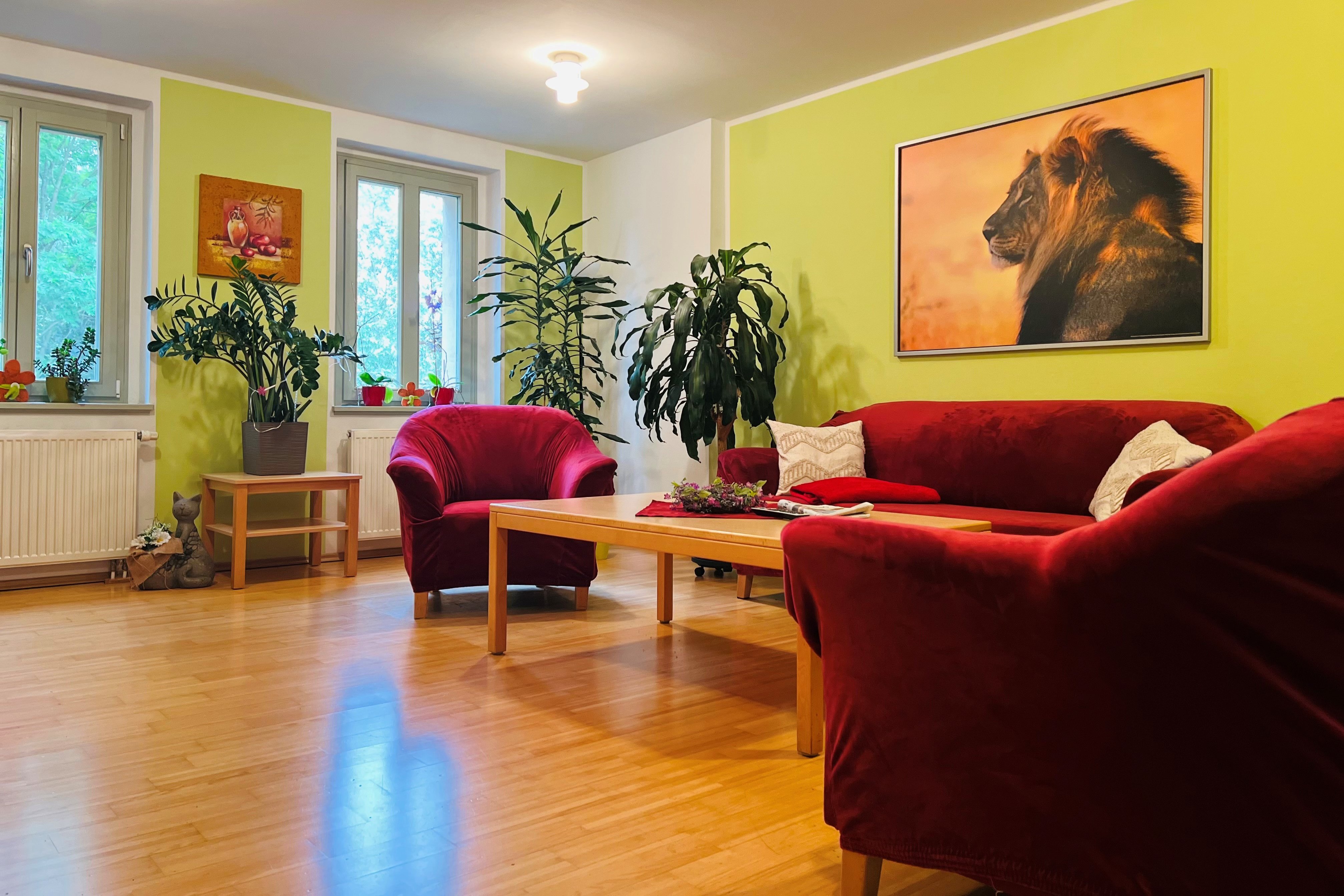 Das Bild zeigt ein Wohnzimmer mit roter Couchgarnitur und grünenwänden. An der Wand hängt das Bild eines Löwen. Grünpflanzen befinden sich im Hintergrund.  