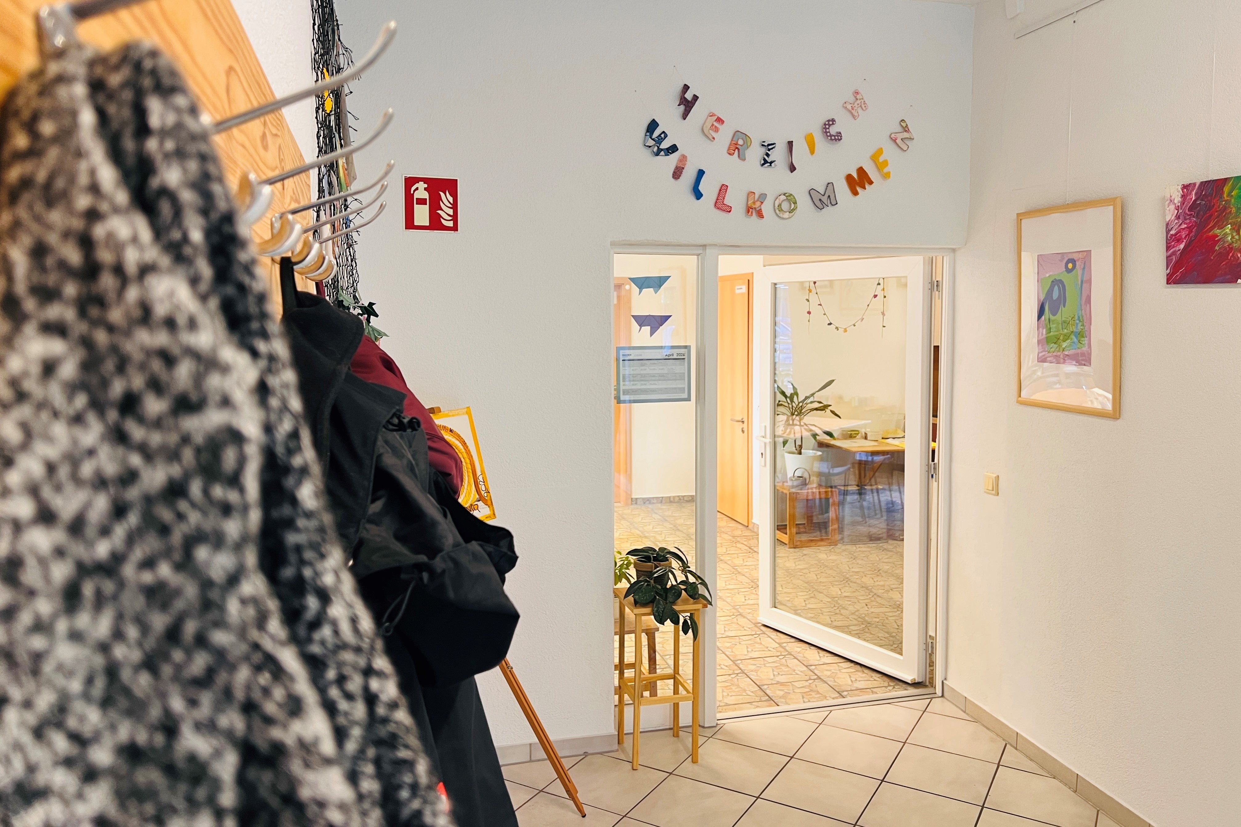 Das Bild zeigt eine Garderobe an der jacken hängen. Im Hintergrund befindet sich die Tür zu einem Raum. Darüber hängen die Worte "Herzlich willkommen"