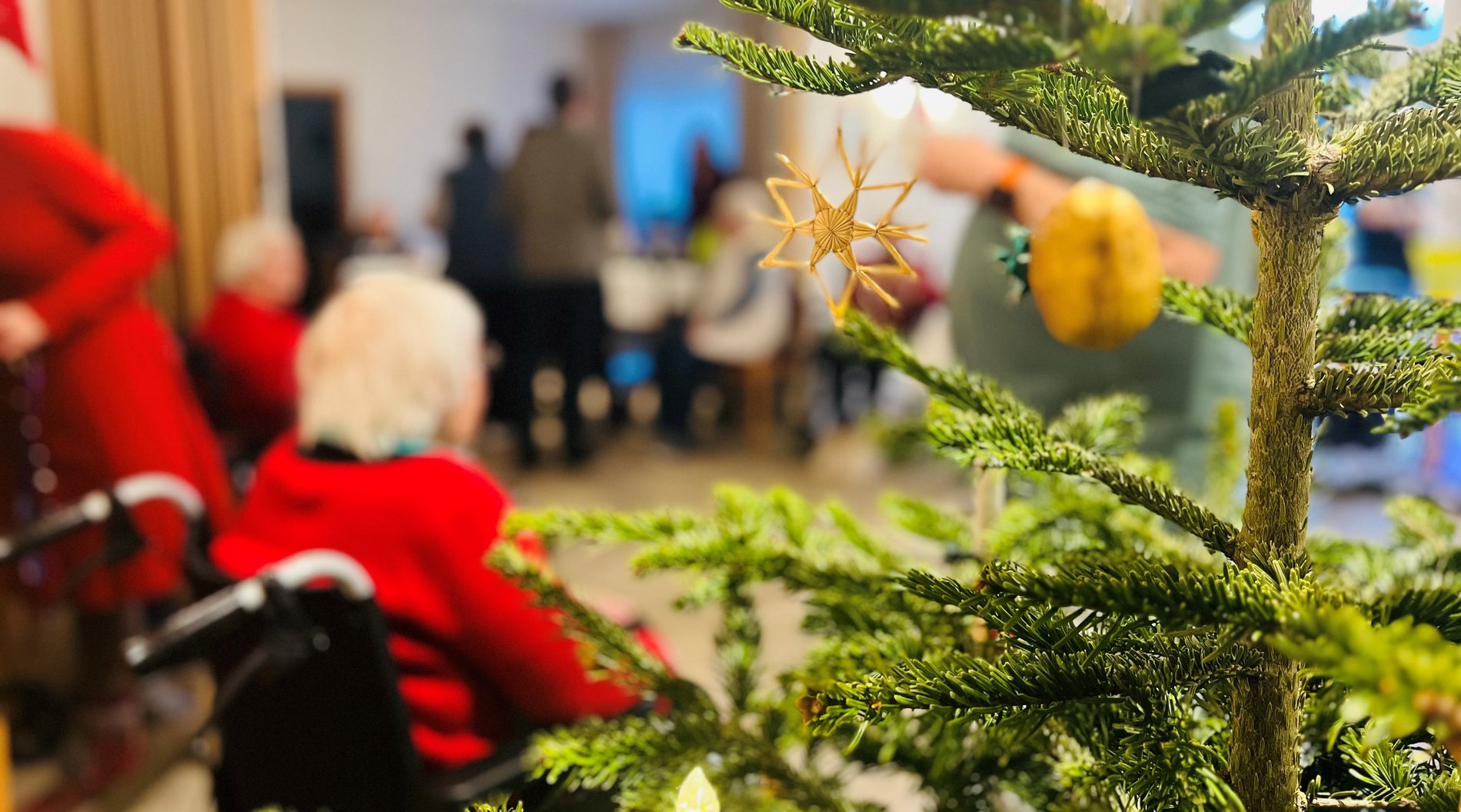 Das Bild zeigt den Weihnachtsmarkt im Saal im Altenpflegeheim Dresden-Plauen. Ein geschmückter Weihnachtsbaum ist im Vordergrund zu sehen. Dahinter sind Bewohner:innen des Altenpflegeheims, die den Weihnachtsmarkt besuchen.