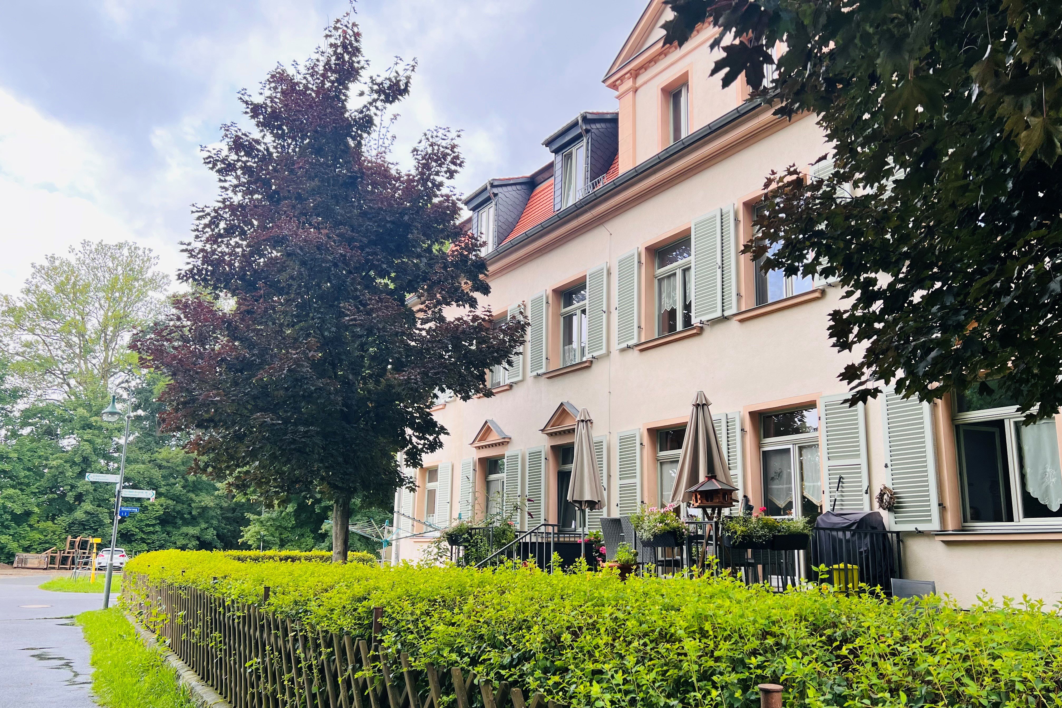 Das Bild zeigt ein ein Haus mit hellgrünen Fensterläden und Terrasse. Eine Hecke mit Holzzaun trennt Straße von Grundstück. 