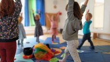 Dieses Bild zeigt Kinder beim Yoga, die die Arme nach oben strecken. In der Mitte sind verschiedene bunte Tücher und eine Klangschale zu sehen.
