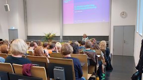 Dieses Bild zeigt den Hörsaal in der Evangelischen Hochschule Dresden, in dem der Abteilungsfachtag stattfand. Das Publikum ist von hinten zu sehen. Vorne steht Suanne Jetter, Abteilungsleiterin, zur Begrüßung.