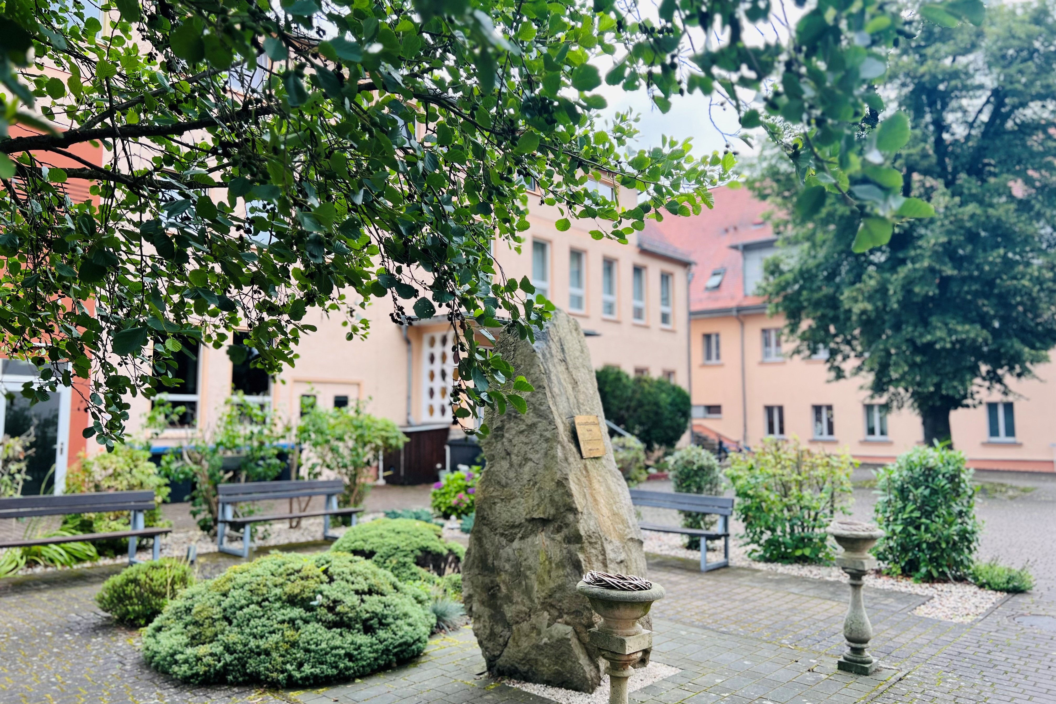 Das Bild zeigt einen gepflasterten Hof. Er ist durch 2 Bäume und mehrere Sträucher begrünt. In der Mitte steht ein großer Sandstein. Drumherum befinden sich mehrere Sitzbänke. 