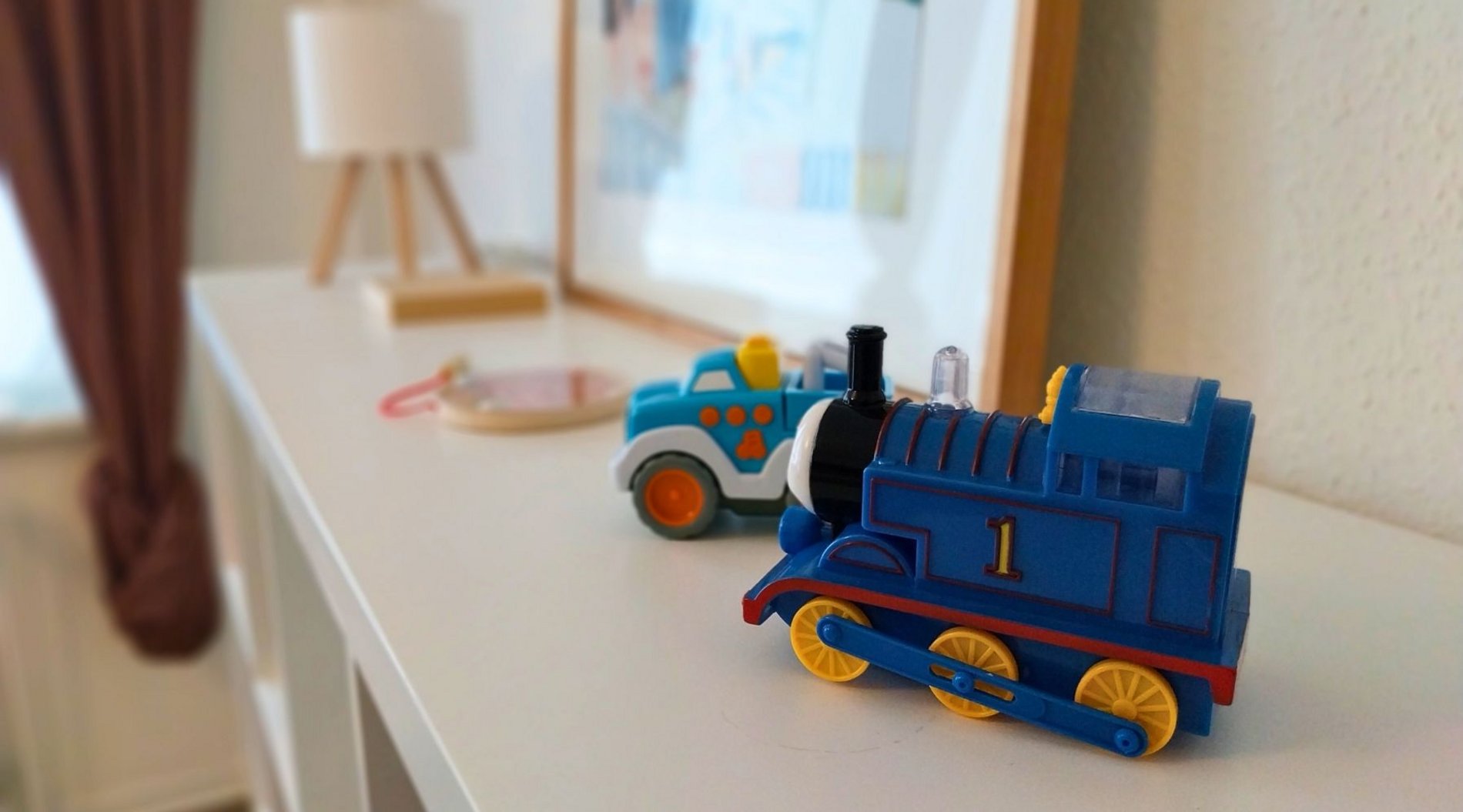 Dies ist eine Nahaufnahme von einem Regal im Wohnzimmer der Wohngruppe Komet. Auf dem Regal stehen eine Lampe, ein Bild sowie Spielzeugfahrzeuge für die Kinder.