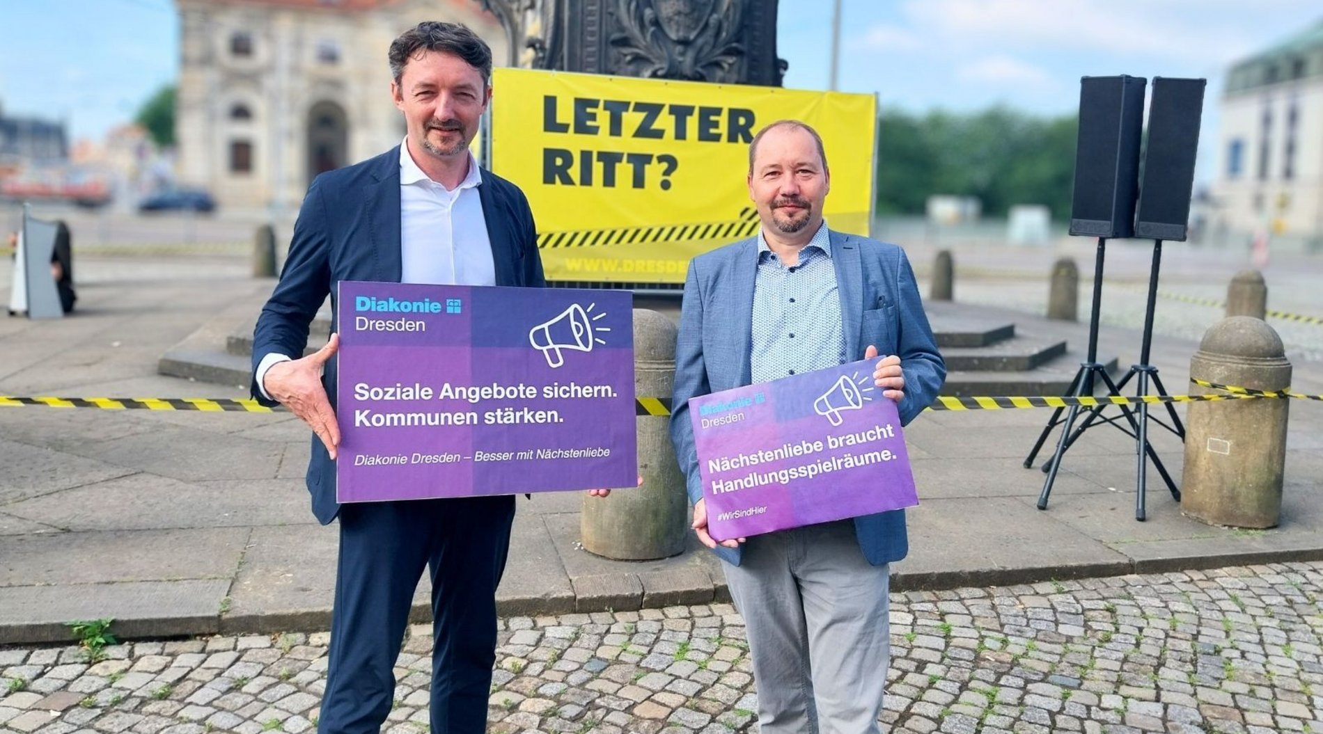 Auf dem Bild sind die beiden Geschäftsführer der Diakonie Dresden, Sven Marschel und Pfarrer Thomas Slesazeck, zu sehen. Sie halten die Diakonie-Schilder "Soziale Angebote sichern. Kommunen stärken." und "Nächstenliebe braucht Handlungsspielräume" in den Händen. Im Hintergrund ist der Sockel des Goldenen Reiters zu sehen mit dem gelben Baller "Letzter Ritt?"