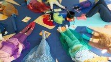 Die Kinder liegen auf diesem Bild im Kreis zur Entspannung auf dem Rücken auf dem Boden und sind jeweils bedeckt von einem bunten Tuch. In der Mitte befindet sich eine Klangschale.
