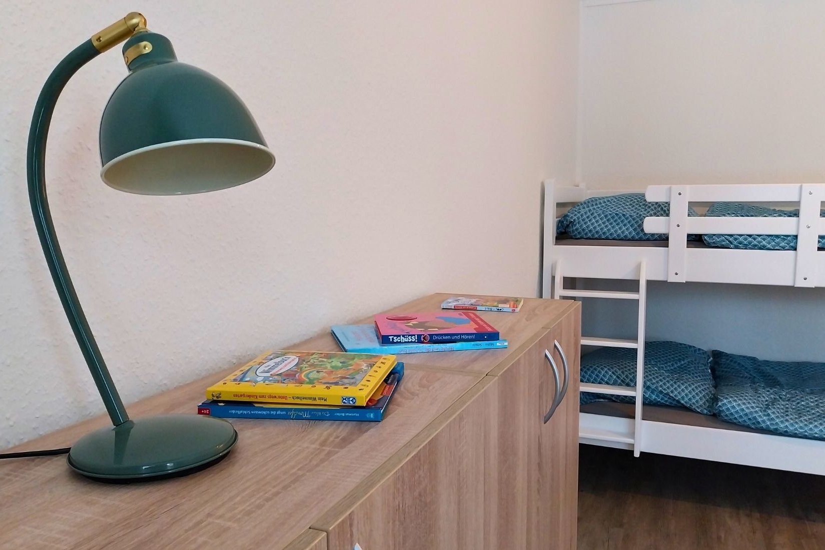Das Bild zeigt ein Zweierzimmer in der Wohngruppe Komet. Ein Hochbett ist zu sehen sowie ein Sideboard, auf dem eine Leselampe steht und verschiedene Bücher liegen.