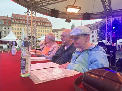 Fünf Personen sitzen auf einer Bühne in Reihe an einem Tisch. Vor ihnen liegen Texte. Die Frau im Vordergrund hat ihre linke Hand auf den Text gelegt und wendet ihren Blick darauf..  