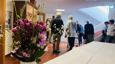 Das Bild zeigt einen Blumenstrauß, im Hintergrund befinden sich Menschen, die miteinander im Gespräch sind.