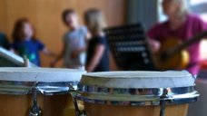 Auch auf diesem Bild sind im Vordergrund Percussions-Instrumente zu sehen. Im Hintergrund spielt eine Pädagogin Gitarre und drei Kinder bewegen sich zur Musik.