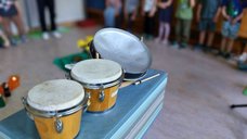 Dieses Bild zeigt im Vordergrund erneut die Percussions-Instrumente, dieses Mal jedoch aus einer anderen Richtung. Im Hintergrund sind viele Kinder im Halbkreis aufgestellt. Die Gesichter der Kinder sind nicht zu erkennen.