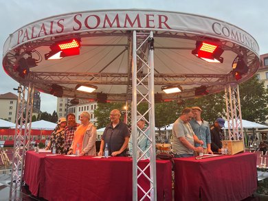 Eine Gruppe Menschen steht in Reihe auf der runden Bühne des Palais Sommer Festivals auf dem Dresdner Neumarkt. Vor ihnen stehen Tische mit Textblättern und Wasserflaschen. Die Personen wirken freudig, gelöst und lächeln. 