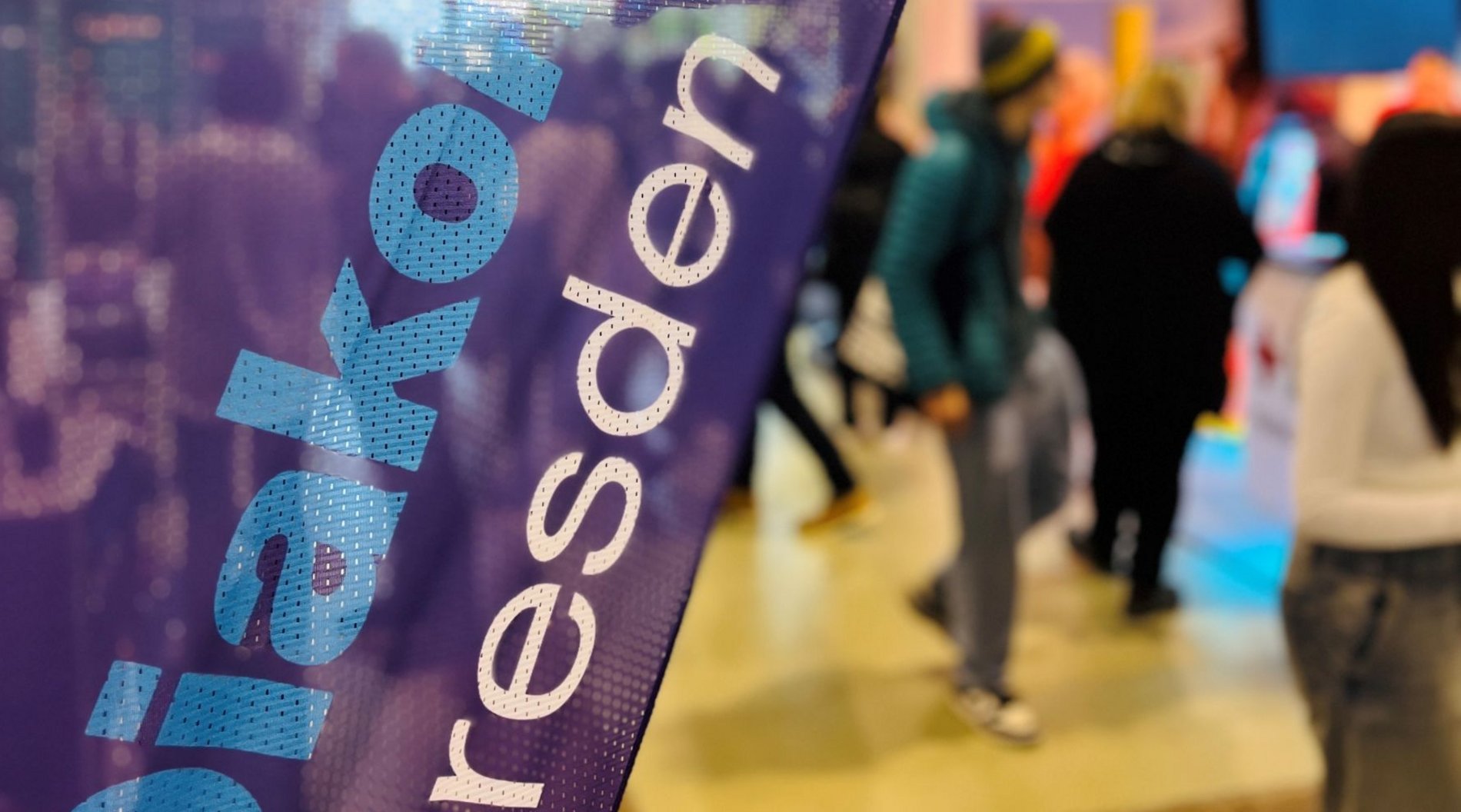 Auf dem Foto ist im Vordergrund eine Werbeflagge der Diakonie Dresden zu sehen. Im Hintergrund bewegen sich Menschen auf dem Messegelände.