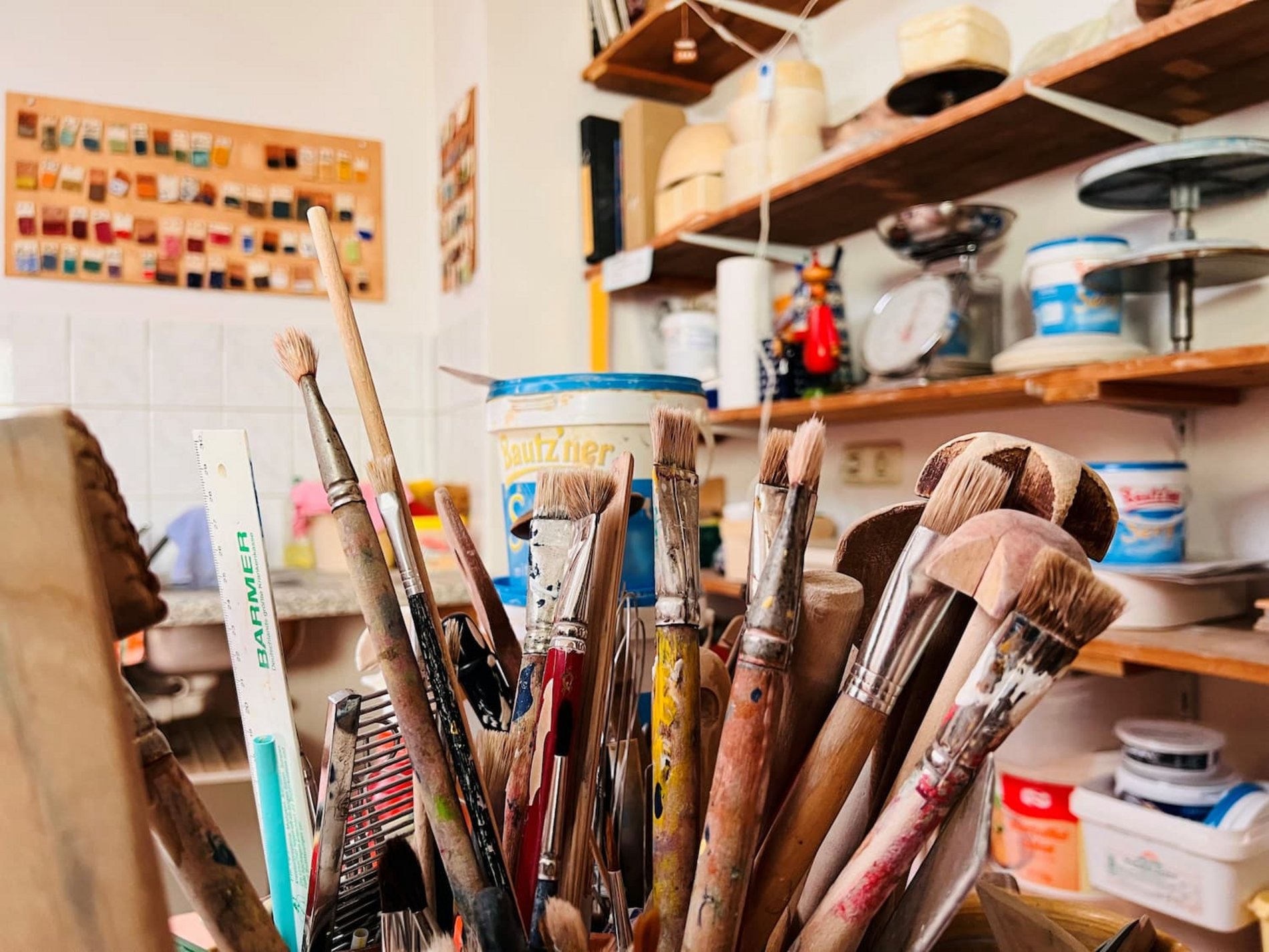 Das Bild zeigt einen Raum, in dem sich unter anderem ein Regal mit verschiedenen Arbeitsmaterialien befinden. Im Vordergrund sind Pinsel zu erkennen. 