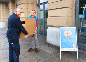 Patrick Büttner von der Bahnhofsmission Dresden und ein Mitarbeiter der Deutschen Bahn tragen einen mit gespendeter Kleidung gefüllten Karton in die Bahnhofsmission Dresden.