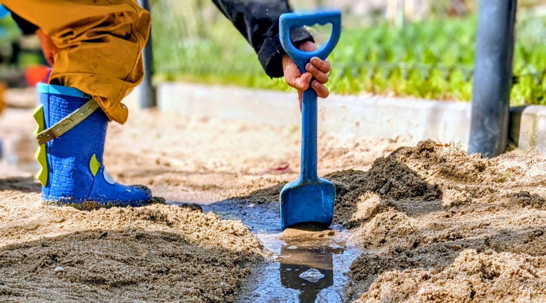 Auf diesem Bild ist ein Kind in Gummistiefeln zu sehen, dass im Sand eine Flutrinne gegraben hat, durch die nun das Wasser läuft.
