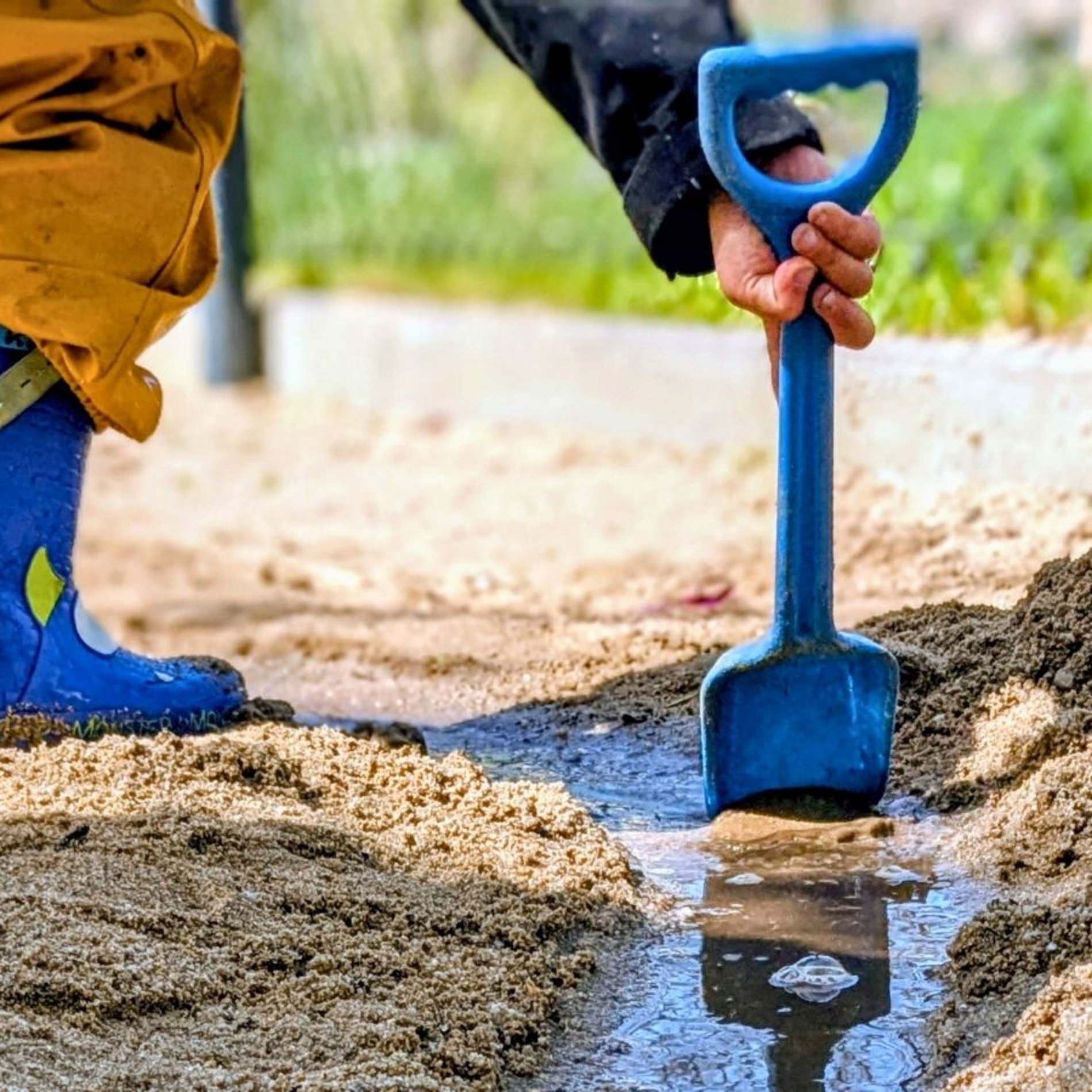 Auf diesem Bild ist ein Kind in Gummistiefeln zu sehen, dass im Sand eine Flutrinne gegraben hat, durch die nun das Wasser läuft.
