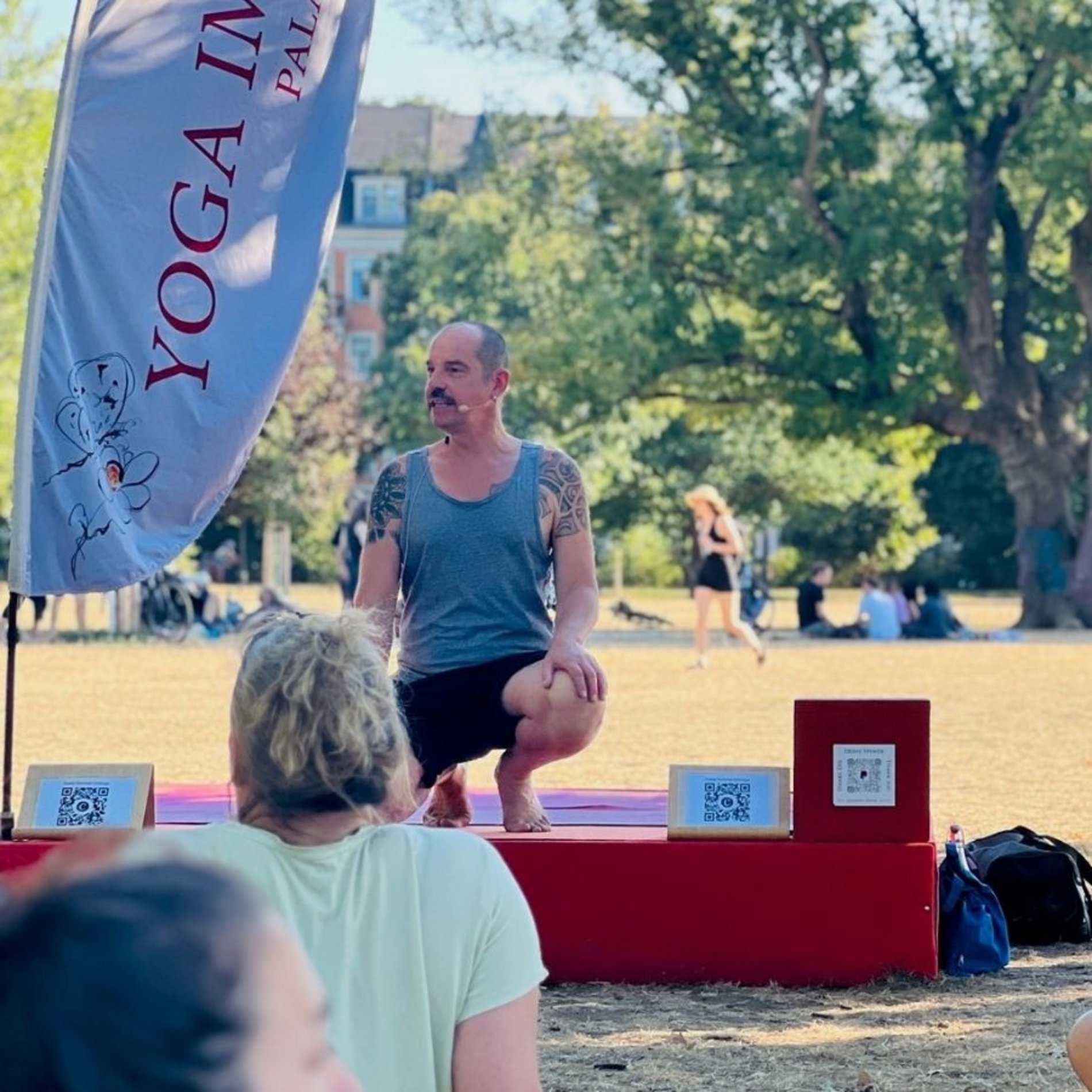 Auf diesem Bild sind Thomas Schwalbe, der die inklusive Yoga-Stunde leitete, sowie die Gebärdendolmetscherin von vorne zu sehen. Von hinten erkennt man Teilnehmende der inklusiven Yoga-Stunde, wie sie auf ihren Matten auf der Wiese des Dresdner Alaunparks sitzen.