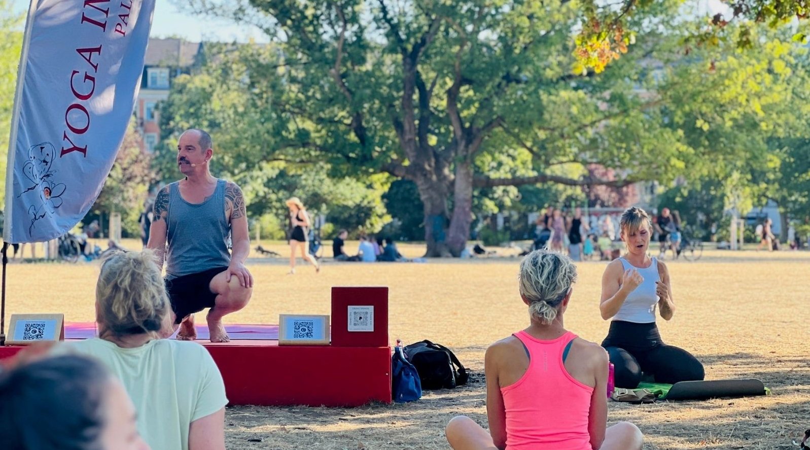 Auf diesem Bild sind Thomas Schwalbe, der die inklusive Yoga-Stunde leitete, sowie die Gebärdendolmetscherin von vorne zu sehen. Von hinten erkennt man Teilnehmende der inklusiven Yoga-Stunde, wie sie auf ihren Matten auf der Wiese des Dresdner Alaunparks sitzen.
