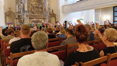 Das Bild zeigt einen Kirchraum mit sitzenden Menschen. Sie wedeln mit farbigen Bändern.