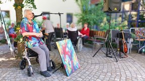 Dieses Bild zeigt Heimbewohner Kurt Behling mit einem seiner Bilder, wie er gerade ins Mikrofon spricht und über seine Kunst und seine Ausstellung berichtet. Sein Hemd trägt das selbe Muster wie das Gemälde vor ihm. Im Hintergrund sitzen andere Bewohner:innen am Tisch und hören seiner Rede zu.