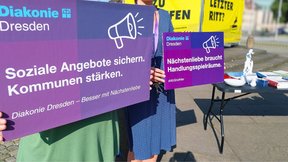 Auf diesem Bild stehen zwei Mitarbeiterinnen der Diakonie Dresden. Ihre Gesichter sind nicht zu erkennen. Sie halten Schilder in den Diakonie-Farben in der Hand, auf denen "Soziale Angebote sichern. Kommunen stärken." und "Nächstenliebe braucht Handlungsspielräume" steht. Im Hintergrund ist das gelbe Banner "Letzter Ritt?" am Goldenen Reiter zu sehen.