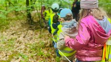 Das Bild zeigt Kinder, die in einem Wald hintereinander an einem Seil entlang gehen. 