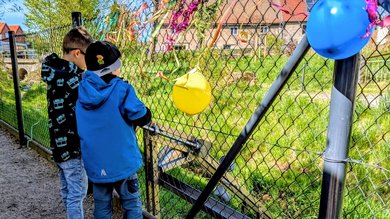 Zwei Kinder stehen am Zaun der Kita und begutachten die neue Schöpfanlage. Am Zaun hängen noch bunte Luftballons von der Einweihung der Anlage.