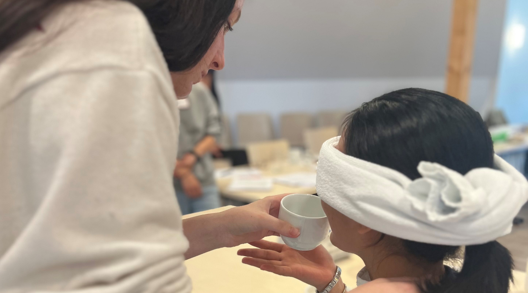 Auf dem Bild ist zu sehen, wie eine stehende Frau einer sitzenden Frau eine Tasse in Mundnähe reicht. Dabei hat die sitzende Frau die Augen mit einem Tuch verbunden.