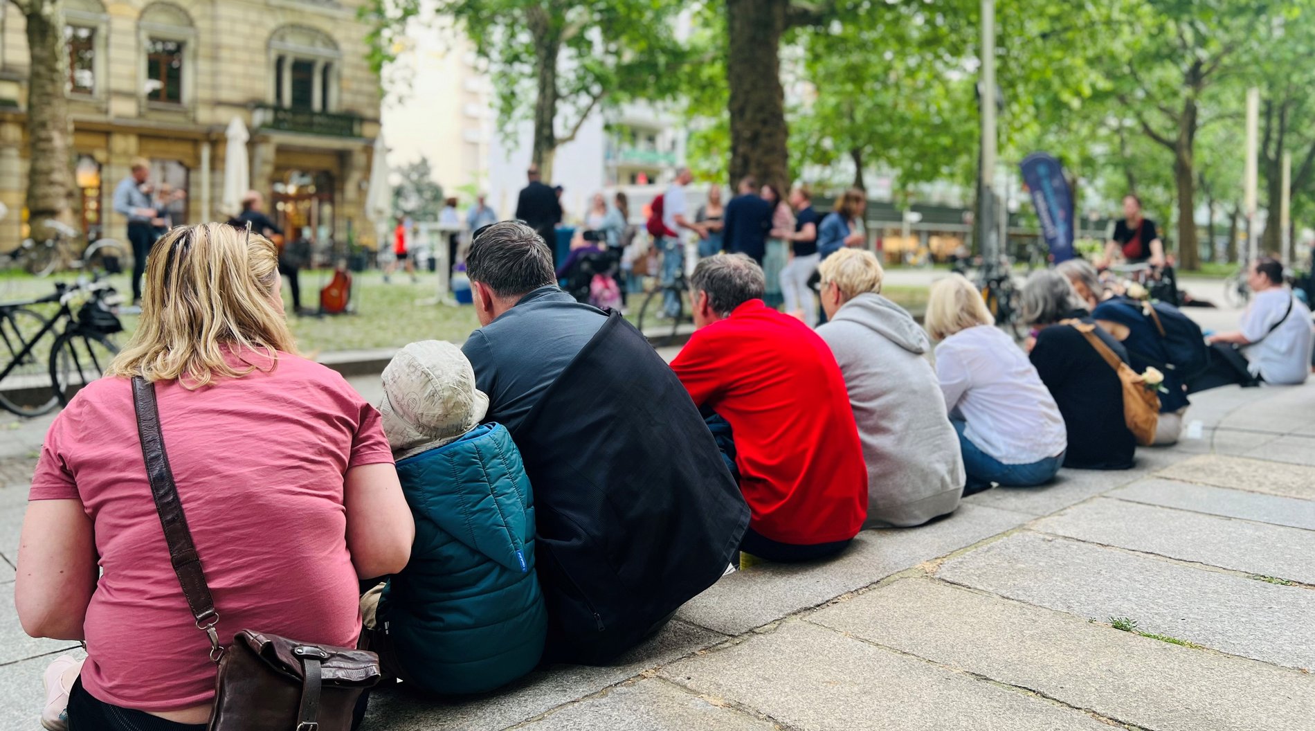 Das Bild zeigt Personen, die auf einer Treppenstufe sitzen und Musikern auf der Straße zuhören.