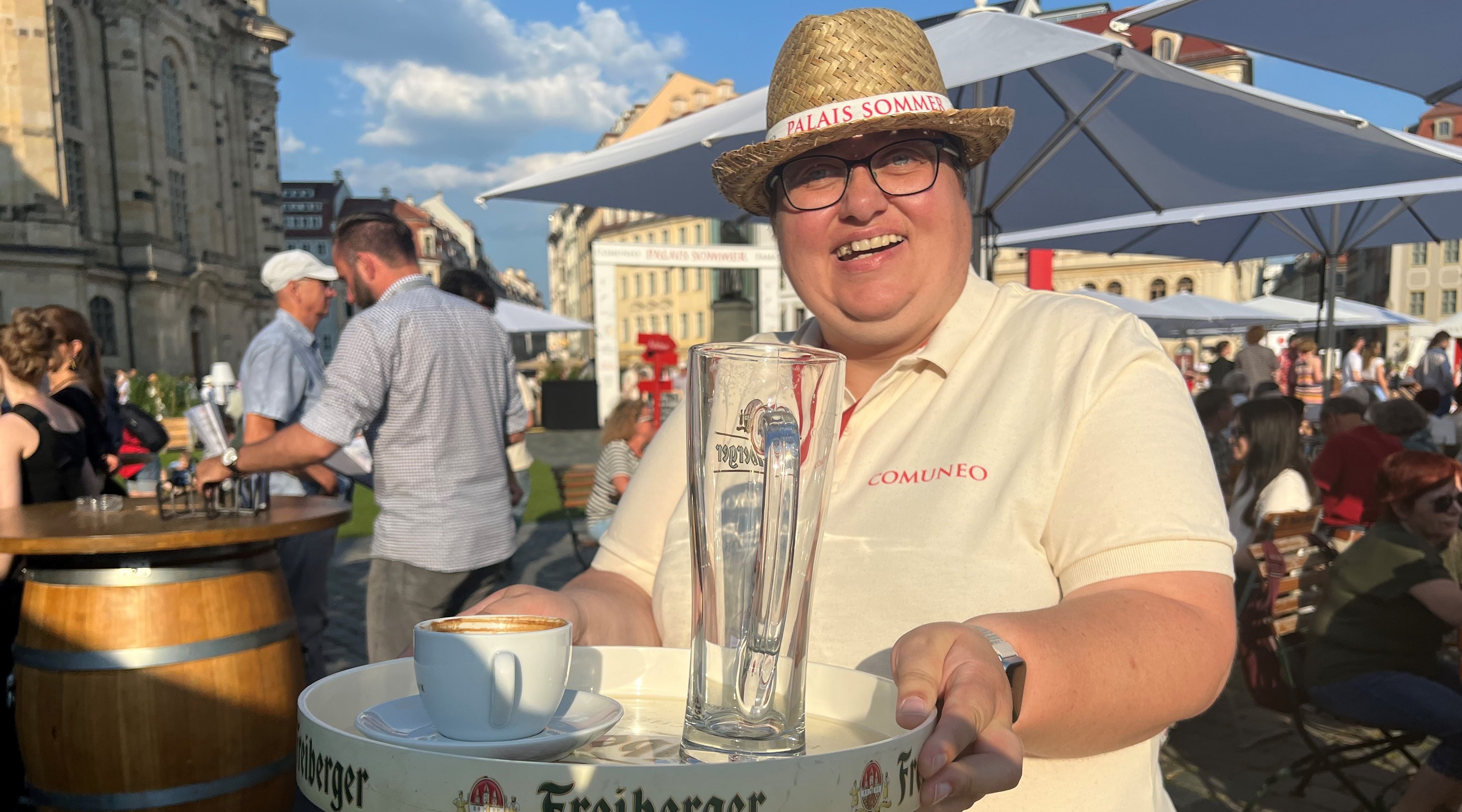 Eine Beschäftigte der Wichern-Werkstätten hält ein Tablett in der Hand. Darauf stehen eine leere Kaffeetasse und ein leeres Bierglas. Sie trägt einen Strohhut und lächelt freundlich in die Kamera
