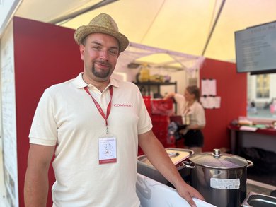 Das Bild zeigt einen Mann mit Stroh-Hut. Er steht vor einem Pavillon, in dem Essen zubereitet wird. Er stützt sich mit seiner linken Hand auf die hüfthohe Begrenzung des Pavillons. Hinter der Begrenzung stehen ein Topf und Speisebehälter.