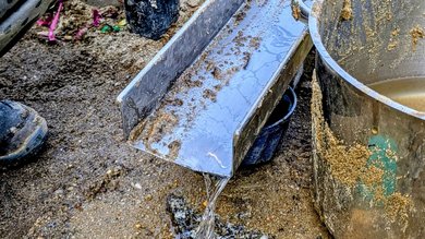 Das Bild zeigt das Ende einer Metallrampe, durch die das Wasser aus dem Bach nun aufs Kitagelände fließt und von Kindern mit Eimern aufgefangen werden kann.