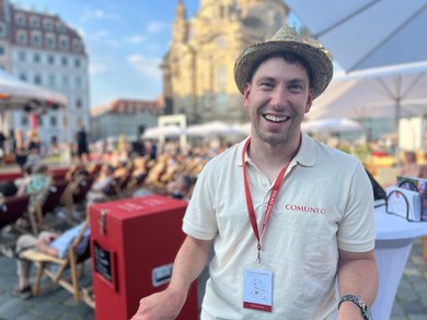 Ein Mann mit Strohhut auf dem Kopf lächelt in die Kamera. Im Hintergrund sind Menschen in Liegestühlen und die Dresdner Frauenkirche zu sehen. Es ist sonnig. 