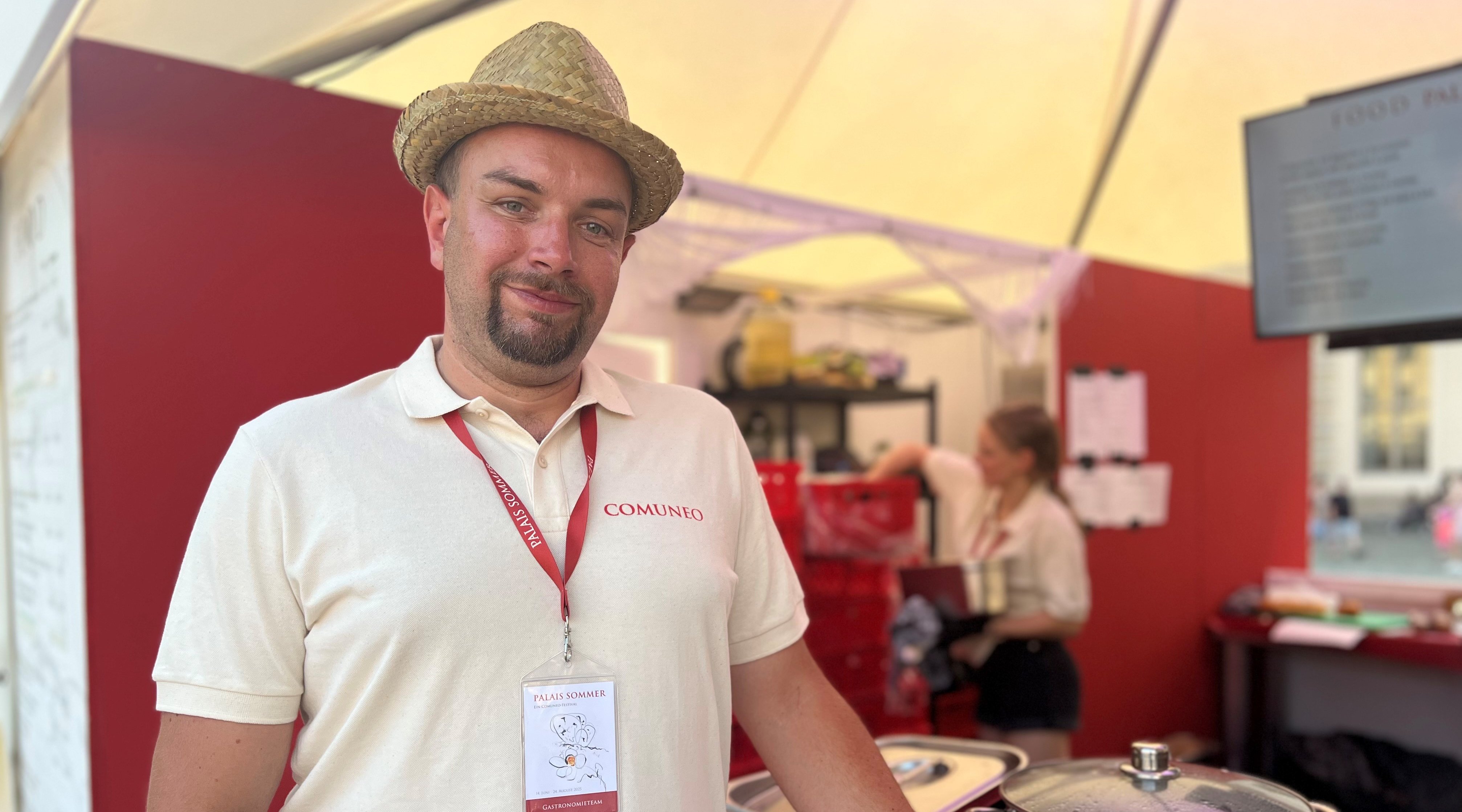 Ein Beschäftigter der Wichern-Werkstätten steht im Cateringbereich des Palais Sommer Festivals und lächelt in die Kamera. Neben ihm stehen Töpfe. Im Hintergrund ist Servicepersonal zu sehen. 