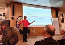 Dieses Foto zeigt die Autorin Melanie Wolfers auf der Bühne während ihres Vortrags. Im Hintergrund ist die Präsentation zu sehen mit den "Quellen der Zuversicht".